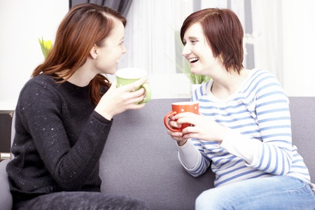 Two happy young female friends with coffee cups enjoying a conversation in the living room at homeの写真素材