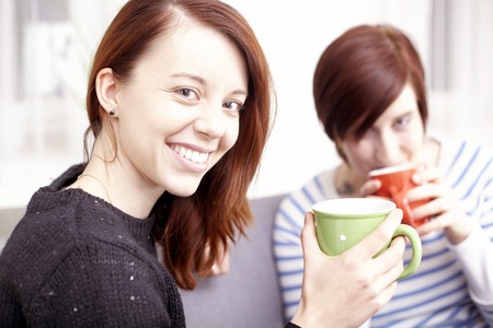 Two happy young female friends with coffee cups enjoying a conversation in the living room at homeの写真素材