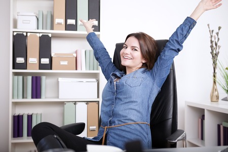 Happy Office Woman in Denim Blouse Sitting on her Office Chair While Extending her Arms and Looking at the Camera.の写真素材