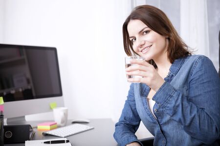 Close up Office Woman Holding a Glass of Water, Leaning on her Desk While looking at the Camera.の写真素材