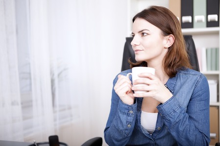 Businesswoman sitting thinking at her desk as she enjoys a coffee break staring off to the side with a quiet smile, with copyspaceの写真素材