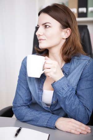 Observant young businesswoman drinking coffee at her desk with the mug in her hands staring off to the side with an attentive expressionの写真素材