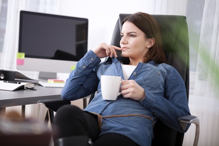 Businesswoman sitting pondering relaxing over a mug of coffee at her desk staring into the distance with a contemplative expressionの写真素材