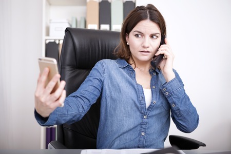 Businesswoman talking on the land line telephone phone in the office while simultaneously checking her mobile with a worried expressionの写真素材