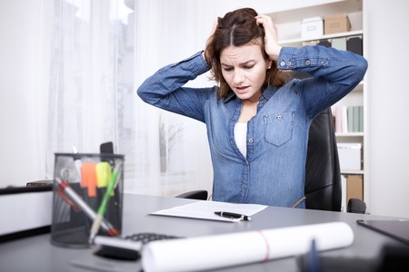 Stressed businesswoman under pressure sitting holding her head with her hands with an anguished expression as she eyes unfinished paperwork on her deskの写真素材