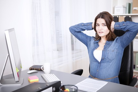 Stressed overworked young businesswoman sitting at her desk with her hands raised to her head and an anguished expressionの写真素材