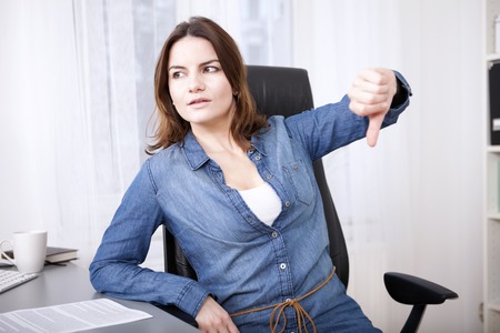 Businesswoman sitting at her desk in the office giving a thumbs down gesture with a serious expression indicating, failure, a negative, voting something down or disapprovalの写真素材