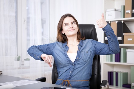 Close up Thoughtful Young Office Woman Sitting on her Chair Showing Thumbs Up and Down Hand Signs While Looking Up.の写真素材