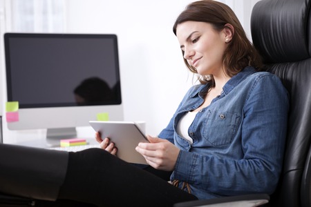 Puzzled businesswoman sitting in her office chair studying her tablet screen with her head tilted to the side and a serious expressionの写真素材