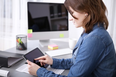 Side view of an attractive young businesswoman surfing the internet on her tablet computer as she sits at the desk using her finger to navigate the touchscreenの写真素材