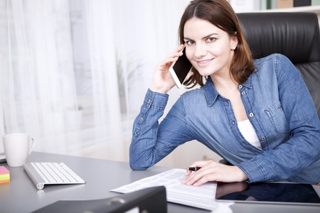 Friendly businesswoman smiling at the camera as she takes a phone call in the office while sitting working at her deskの写真素材