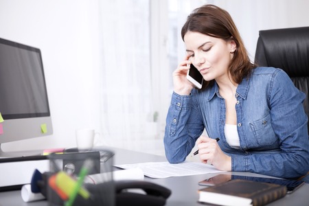 Serious businesswoman listening to a telephonephone call at her desk reading peperwork in front of her as she listensの写真素材