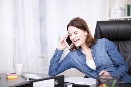 Close up Very Happy Office Woman Laughing While Talking on the Phone While Leaning at her Worktable.の写真素材