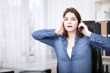 Businesswoman reacting in shock to a phone call sitting at her dek with a wide eyed expression and her hand to her faceの写真素材