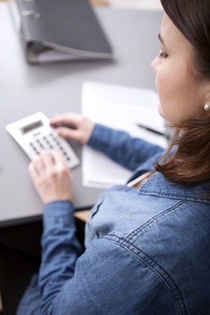 Over the shoulder view from behind of a businesswoman doing calculations at her desk on a desktop calculator as she compiles a business reportの写真素材