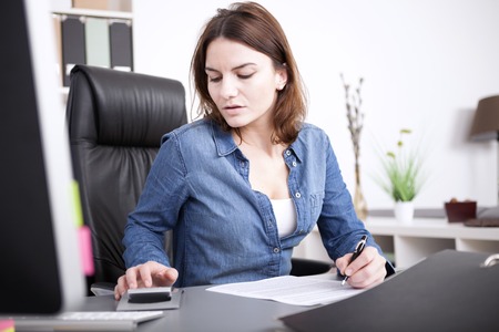 Businesswoman checking figures on a calculator as she sits at her desk in the office proof reading a report and making notesの写真素材