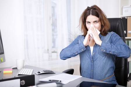 Young businesswoman with dishevelled hair with a cold and flu sitting at her desk in the office blowing her nose on a tissueの写真素材