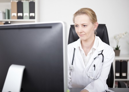Serious Female Physician with Stethoscope on her Shoulders Looking at the Computer Monitor on her Table While Checking Some Information.の写真素材