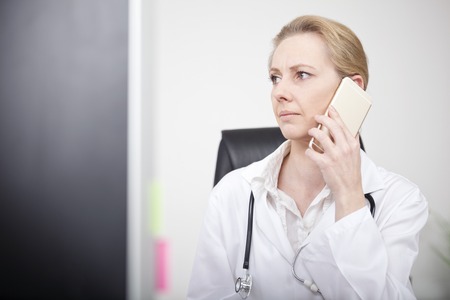 Close up Pensive Blond Woman Doctor Calling to Someone on Mobile Phone While Looking to Far Left Seriouslyの写真素材