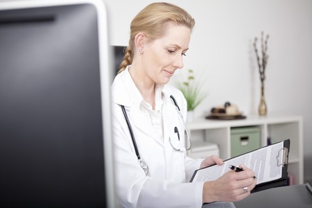 Smiling Adult Female Doctor at her Office Writing Diagnosis on a Paper Attached to a Clipping Board.の写真素材