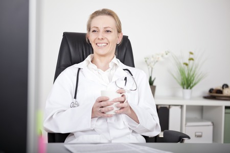 Happy Adult Female Doctor Relaxing on her Chair at the Office, Holding a Cup of Coffee While Looking Afar.の写真素材