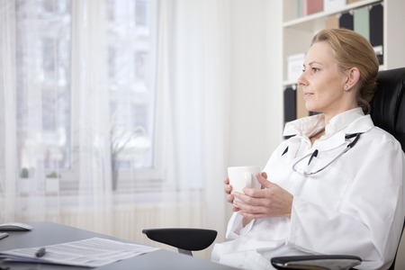 Side View Shot of a Thoughtful Medical Doctor Holding a Cup of Drink While Sitting on a Chair at her Office and Looking Afar.の写真素材