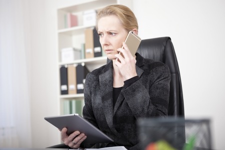 Close up Pensive Manageress in Black Business Attire Holding a Tablet Device While Calling to Someone Using Mobile Phone and Looking to the Left.の写真素材