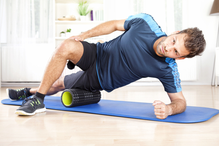 Handsome Athletic Young Man Doing Side Planking with Foam Roller on his Thigh.の写真素材