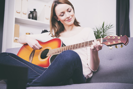 Pretty young girl having her legs crossed playing some record on guitar sitting on couchの写真素材