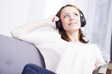 Attractive young girl enjoys listening to music with headphones on while sitting lazy on sofaの写真素材