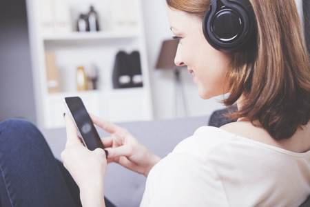 Beautiful brown haired girl sitting on sofa with smartphone in her hands enjoying lisenting to music wearing headphonesの写真素材