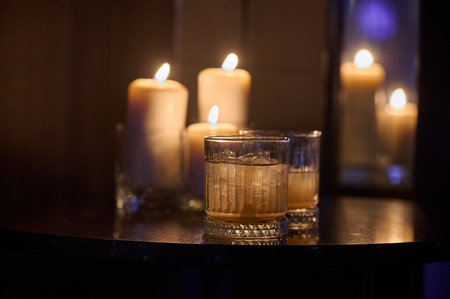 Two whiskey glasses on reflective table with blurred candlelight warm ambient moody low-light horizontal close-up commercial bar decor shotの写真素材