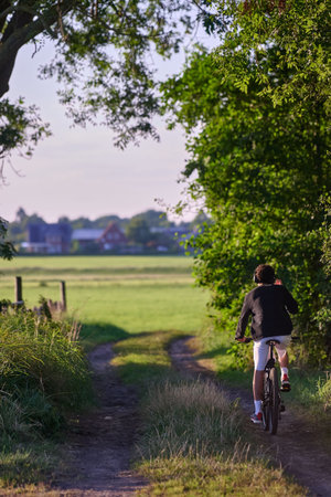 Rear view cyclist biking dirt path green meadow countryside farmhouse background summer outdoor Germany vertical active recreationの写真素材