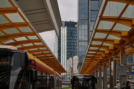 City bus under yellow geometric glass canopy, urban transit hub, modern architecture, tall office buildings, downtown street view, perspective composition, contemporary designの写真素材