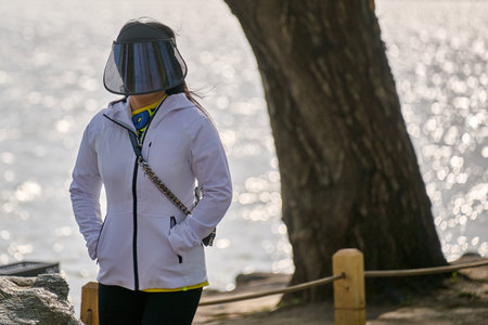 Woman standing contemplatively at waterside wearing white protective jacket and visor, gazing at...