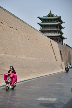 Woman on bright pink scooter passing monumental stone wall and pagoda tower, Pingyao ancient city China, dramatic architectural contrastのeditorial素材