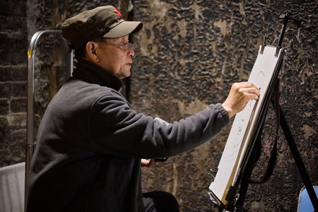 Senior Asian man in white cap painting on canvas in alley, street artist, traditional architecture backdrop, horizontal orientation, daytime light, occupation focus, creativity and craftsmanship, suitable for cultural documentation and artistic pursuits.のeditorial素材