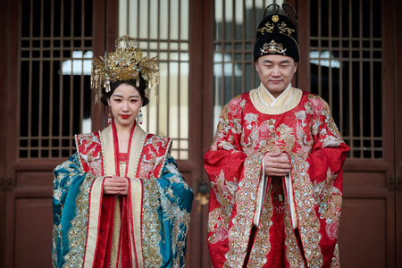 Couple in traditional Hanfu imperial wedding attire, ornate golden crowns, embroidered red and teal robes, temple building backgroundのeditorial素材