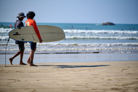 Two men walking along the sandy shore carrying a surfboard at Weligama Beach, Matara District,...