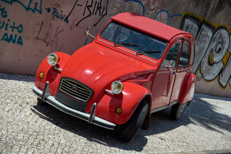 Red vintage Citroen 2CV parked on a cobblestone street against a graffiti wall, Lisbon, Portugal,...