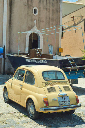 Vertical shot of a classic yellow Fiat 500 and a dry-docked fishing boat in a stone plaza near a...