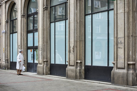 Senior woman with walking stick and plastic rain bonnet standing on the sidewalk by a classical stone building, Krakow, Poland, Europeのeditorial素材