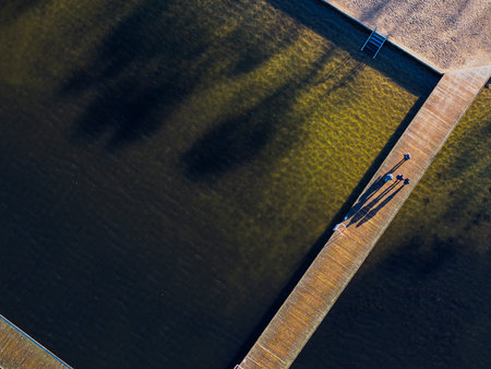 Aerial view of the long shadows of three people walking on a wooden pier extending from the sandy shore into Ukiel Lake, Olsztyn, Warmian-Masurian, Poland, Europe.のeditorial素材