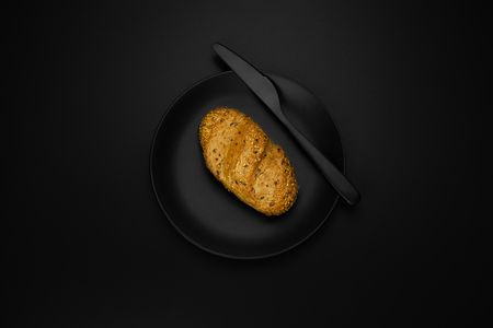 A roll on a black plate with a knife and salt shaker on a black background, shot from aboveの写真素材