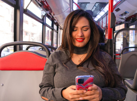 Young Latina woman sitting and smiling on a bus while typing on her cell phone, image where the...