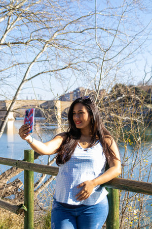 Young pregnant woman taking a selfie by the Ebro river in Zaragoza, Spain. She is wearing blue clothes and sunglassesの写真素材