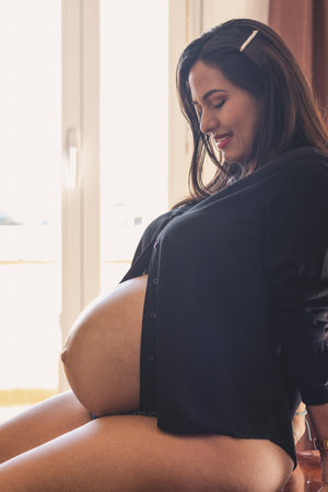 Young pregnant woman, sitting looking at her belly with a window behind and black blouse.の写真素材