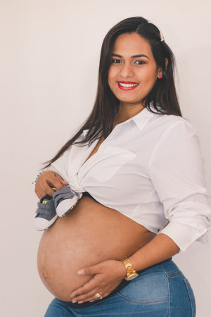 Pregnant young latin woman smiling while holding small shoes on her bellyの写真素材