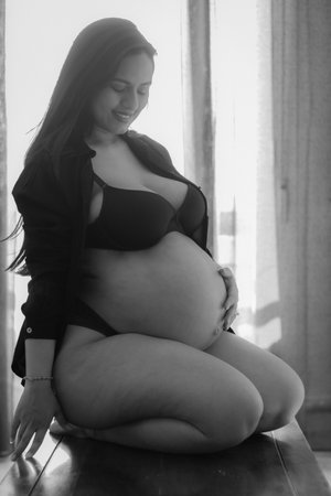 Young pregnant Latina woman on her knees touching her belly, wearing black underwear with a window in the background. Black and white photoの写真素材