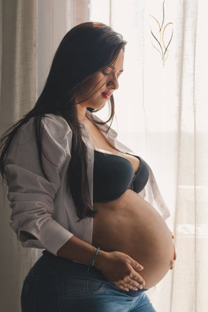 Pregnant latin woman with her hands on her belly, smiling next to a window, wearing a white blouse and black bra.の写真素材